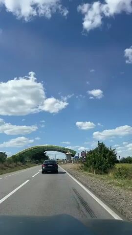 A black car drives down a paved road under a bright blue sky with white clouds. The car is approaching an arched structure that spans the road. The archway is decorated with green and yellow elements. A small building or tower is situated to the right of the road under the archway. There is a person on a ladder on top of this structure, appearing to be working. Road signs are visible on the right side of the road. Trees and vegetation line the sides of the road. The view is from inside another vehicle, looking forward through its windshield.