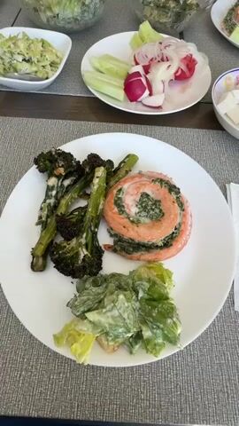 A top-down shot shows a white plate with a meal of roasted broccoli, a salmon roulade, and a small side salad. The salmon roulade is spiraled and filled with a green mixture, possibly spinach and cheese. The broccoli appears to be roasted with a light sauce. The salad consists of lettuce leaves tossed in a creamy dressing. In the background, other plates with food are visible, including a plate with sliced radishes and cucumbers, and a bowl of what looks like a coleslaw or pasta salad. The table setting includes a gray placemat. The lighting is natural and appears to be from an overhead source, casting soft shadows. The overall scene suggests a home-cooked meal.