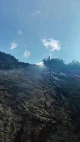 A low-angle shot pans up a steep, rocky mountainside. Sunlight streams down from the top left, illuminating a patch of trees and a thin waterfall cascading down the rock face. The sky above is blue with scattered white clouds.