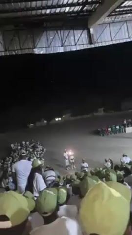 A video shows a large group of people in matching white shirts and light green caps. Some are sitting in bleachers under a roof, facing a large open field. In the distance, there is a crowd of people in mixed colors, and two individuals wearing bright white shirts stand facing each other with a light between them. The scene is illuminated by overhead lights.