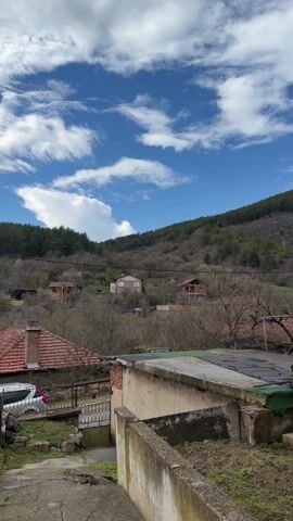 The camera is focused on a rural setting, seemingly a small village nestled at the base of a hill or mountain covered with trees. The sky is blue with scattered white clouds, although some darker clouds are present as well. In the foreground, there is a rough concrete structure with an uneven surface, possibly a wall or part of a building. Beyond this, the scene opens up to reveal a cluster of houses with red-tiled roofs. A white car is parked near one of the houses. Overhead, a power line runs through the frame. The overall impression is of a quiet, rural landscape with a mix of natural elements and human habitation.