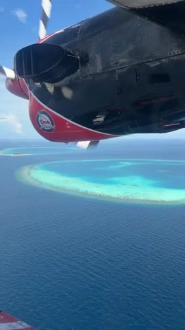 A close-up, aerial shot shows the underside of a twin-engine seaplane as it flies over a tropical ocean. The plane has black and red paint with a logo on the red section of the engine cowling. The propeller is blurred due to its rapid rotation. Below the plane, a circular reef or atoll is visible, with turquoise water inside the reef and deeper blue water outside. The sky is clear and blue with some scattered clouds. The video is very short, lasting only a few seconds.