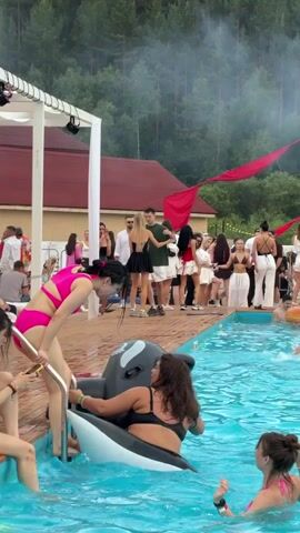This video captures a lively pool party scene at an outdoor venue in Дрокино, Russia. The central focus is a bright blue swimming pool filled with people enjoying themselves. In the foreground, two women are interacting with a large inflatable killer whale. One woman, wearing a vibrant pink bikini, is climbing out of the pool, holding onto the inflatable. Another woman, in a black bikini, is already seated on the killer whale.

The background reveals a gathering of people on a wooden deck surrounding the pool. Many are dressed in summer attire, with some in swimwear and others in fashionable outfits. A white structure, possibly a canopy or stage, is visible on the left, adorned with lights. Red fabric banners are draped across the scene, adding a festive touch. Beyond the deck, a building with a red roof is situated, and behind that, a dense forest covers a hillside, suggesting a natural, somewhat remote setting.

The atmosphere appears to be one of celebration and leisure. People are seen chatting, standing around, and a few are in the pool, some with inflatable rings. The lighting suggests it is daytime, likely late afternoon or early evening, given the soft light. The weather seems pleasant for an outdoor event. The overall mood is energetic and social, with a clear emphasis on fun and relaxation. No visible text is present in the video.
