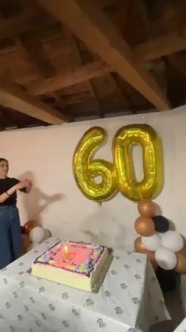 Here is a description of the video:
The video shows a birthday celebration, presumably for someone turning 60. The scene is indoors with exposed wooden beams on the ceiling. A table is set up with a decorated birthday cake with a lit candle, and two large gold foil balloons forming the number "60" are attached to the wall behind it. A balloon arrangement in gold, black, and white is also next to the "60" balloons. Initially, a person is standing near the table on the left side of the screen, and the camera focuses on the cake and decorations. The camera then pans up to show a man standing on what appears to be a table against the wall. He has his arms raised, seemingly in a celebratory pose. There's a speaker and a bottle of green liquid on the table to his side. The camera then pans back down to show the cake and the "60" balloons again. The scene appears to be a casual, intimate birthday gathering.