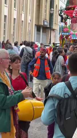 A man wearing a green jacket and yellow garment stands to the left, holding a small drum and a stick. He is looking towards the right and smiling. Beside him, a woman in a red garment also looks to the right. Behind them, a crowd of people are walking on a street. Some individuals are wearing high-visibility vests. The buildings in the background are light-colored and appear to be shops or offices.