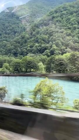 The media file captures a scenic view of a mountainous landscape with a turquoise river flowing through it. Lush green trees and dense foliage cover the slopes of the mountains, which recede into the distance under a cloudy sky. The perspective suggests the view is from a moving vehicle, possibly a train or car, as indicated by the motion blur.

In the foreground, along the riverbank, there are several people. Some are gathered near a wooden structure that appears to be a pier or a boardwalk, while others are seen in small groups, possibly relaxing or enjoying the scenery. The presence of people enjoying themselves in this natural setting suggests a recreational or tourist activity. The weather appears to be overcast, with clouds obscuring parts of the sky and mountains. The time of day is likely daytime, given the visible light.

The setting is identified as Bzypta, Abkhazia, which provides a geographical context for the natural beauty depicted. The vibrant turquoise color of the river is a striking element in the scene, contrasting with the surrounding greenery and the muted tones of the sky. The overall impression is one of natural splendor and tranquil recreation, despite the overcast conditions.