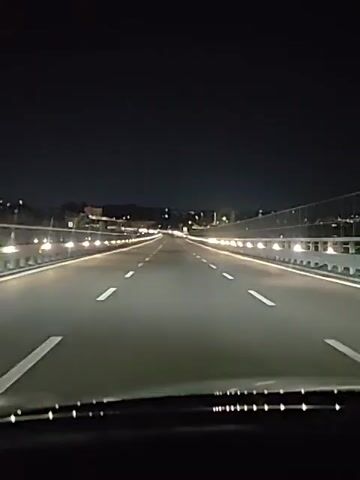 A first-person perspective video shows a car driving on a multi-lane highway at night. The road is well-lit by streetlights and the car's headlights. The lane markings are clearly visible, guiding the car forward. In the distance, city lights and a dark sky are visible. The camera is positioned behind the windshield, capturing the view of the road ahead. The video is short, with the timestamps indicating it spans just a few seconds. The audio includes spoken words, possibly directions or commentary, but the content is not discernible from the provided transcript.
