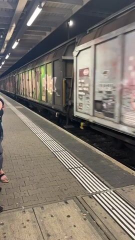 The video shows a train passing through a station platform. The train is composed of several freight cars, many of which are covered in graffiti. The platform is paved with rectangular stones and has white tactile paving stripes along the edge. Overhead lights illuminate the station. A person is partially visible on the left side of the frame, wearing a black t-shirt and denim shorts. The train appears to be moving at a moderate speed, indicated by the motion blur on the graffiti. Some of the graffiti is in green, pink, orange, and red. There are also some warning symbols and text on the train cars, such as a "no smoking" symbol. The lighting suggests it could be daytime or a well-lit indoor station. The overall scene depicts a functional train station with the added visual element of graffiti-covered cargo.