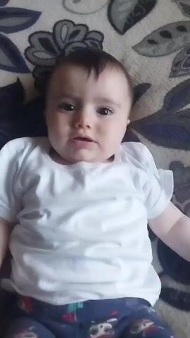 The video shows a close-up, overhead shot of a baby lying on a blanket with a floral pattern. The baby is wearing a white t-shirt and patterned pants. The baby's head is tilted slightly to the right and their gaze is directed at the camera. Their facial expression changes throughout the video, starting with a neutral look, then transitioning to smiles and laughter, and finally returning to a neutral expression. The lighting appears to be natural, coming from above. The background consists of the blanket, which is a neutral gray color with dark blue and white floral motifs. The baby's skin tone is fair, and they have dark hair and brown eyes. Their hands are visible on either side of their body. The video is approximately 5 seconds long.