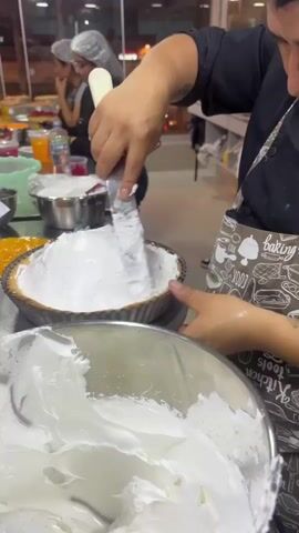A person wearing a black chef's coat and a white hairnet is frosting a pie with a spatula. The pie is in a silver pie pan, and the frosting is white. Another person, also wearing a hairnet, is visible in the background. There are bowls and other baking supplies on a counter. The setting appears to be a bakery or kitchen.