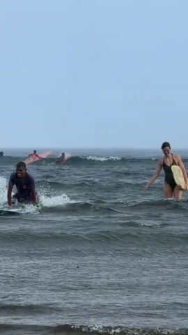 Two individuals are in the ocean with their surfboards. A young man wearing a dark-colored short-sleeved shirt is sitting on his surfboard in the water and faces the camera. A woman wearing a one-piece swimsuit carries her surfboard and walks through the water toward the shore, but she is further out in the water compared to the man. They raise their hands in the air as if high-fiving. Other surfers are visible in the distance. The sky is overcast and the ocean is choppy.