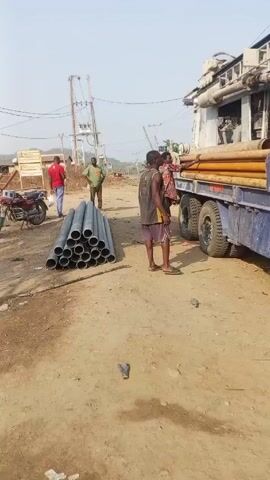 The video shows a group of men working near a drilling rig. The rig is mounted on a blue truck and has several pipes attached to its side. One man is standing on the truck, apparently unloading pipes. Another man is walking toward the truck with a long stick. Several other men stand nearby watching. A pile of gray pipes is on the ground, and a motorcycle is parked near them. The ground is dirt and gravel. In the background is a utility pole and some trees. The sky is clear.