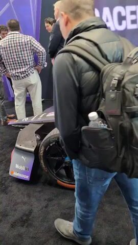 The video shows a purple and black race car on display at an event. People can be seen walking around and looking at the car. The car has several sponsors, including TAG Heuer, AVEVA, Porsche, and TDK. There is a sign next to the car with information about it. The event appears to be taking place indoors on a black carpeted floor. The lighting is bright.