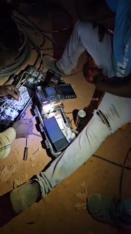 Two people are working on a complex electrical box on a dirt ground at night. One person is kneeling and using their hands to manipulate wires within the box, while the other person is sitting nearby. The electrical box is open and illuminated, revealing intricate wiring and components. Tools, including a screwdriver and a wrench, are scattered around the box. There is also a can of drink near the box. One person is wearing ripped jeans. Cables and other electrical equipment are visible in the background.