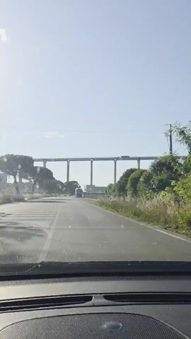 The video is filmed from the perspective of someone in the driver's seat of a car, looking out at a road. The car is moving forward on a paved road with white lane markings and a pedestrian crossing. On the left side of the road, there are trees and a blue sky with sunlight filtering through. On the right side, there is a low stone wall and a building. In the distance, a long elevated structure with multiple pillars spans across the horizon. There are some vehicles on the road, including a truck. The overall environment appears to be a rural or semi-urban area on a bright, clear day.