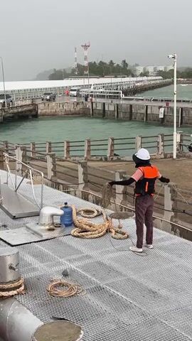 In a coastal setting, likely Don Sak, Thailand, a person stands on a metal-grated deck of a vessel. They are wearing a white hard hat, a black face covering, and an orange life vest over a dark shirt and reddish-brown pants. Their stance and the way they are holding a coiled rope suggest they are preparing to tie up the boat or handle mooring lines.

Behind the person, a pier stretches into the distance, lined with a railing and supporting a roadway with vehicles and a structure that appears to be a terminal building. The water is a vibrant turquoise and appears choppy, indicating a possibly breezy or active sea state. In the background, a bridge connects to what looks like an island or further landmass, with industrial structures and communication towers visible against a hazy sky. The weather appears overcast, possibly signaling an impending rain shower or generally humid conditions typical of a tropical climate.

The deck of the boat itself is utilitarian, with various metal fixtures, including a large white water jug, and several thick ropes coiled neatly. The edge of the boat shows protective tires, weathered and worn, acting as fenders against the pier or other vessels. The overall impression is of a working harbor or ferry terminal, with the focus on the practicalities of maritime operations. The time of day is not explicitly indicated, but the lighting suggests daytime. No visible text is present in the media.