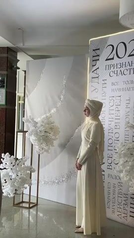 A young woman in a cream-colored hijab and dress stands next to a large white backdrop with gold accents. The backdrop has the year 2026 prominently displayed, along with text in Russian that seems to be motivational phrases. To the left of the woman, a white floral arrangement is displayed on a gold stand. To the right, another white floral arrangement frames the right side of the backdrop. The woman is looking to her left, away from the camera. The environment appears to be an indoor event space, possibly a photo booth or a decorated area for an event.