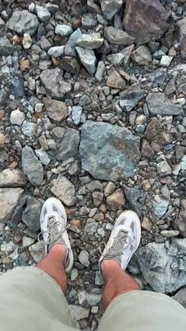 The video opens with a first-person view looking down at a person's feet wearing light-colored athletic shoes on a rocky, uneven ground composed of various shades of grey and brown stones. The camera then slowly tilts upwards, revealing a landscape with green foliage in the midground and a large body of water in the background. Several floating structures, resembling houseboats or docks, are visible on the water. Distant hills or mountains frame the horizon, where a sunset or sunrise casts orange and pink hues across a partly cloudy sky.