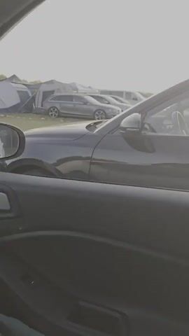 The video shows a parked dark colored car in the foreground. In the background, several other cars are parked. The sun is setting or rising in the sky and is visible through clouds. The scene appears to be an outdoor parking lot.