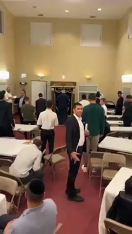 The video depicts an interior space, likely a synagogue or community hall, with a group of men, many of whom appear to be of Jewish faith, present. They are dressed in a variety of attire, including suits, collared shirts, and traditional Jewish garments. The room is furnished with rows of folding chairs and small tables. A man in a black suit and white shirt stands in the foreground, gesturing towards the camera with a smile. In the background, several men are gathered near what appears to be an ark or other religious structure. The overall atmosphere seems to be social and possibly pre or post-event.