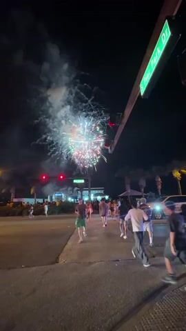 The video shows a street scene at night with fireworks exploding in the sky. Several people are walking on the sidewalk, some are looking at the fireworks. There is a street sign that says "S Pier Park Dr". Traffic lights are visible, one of which is red.