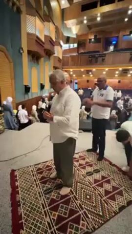 The video shows a group of people, primarily men and boys, standing and praying on prayer rugs inside a large auditorium. The auditorium has tiered seating, and some women in white hijabs can be seen seated in the background. The men are dressed in a mix of casual clothing, with some wearing white shirts and dark pants. Two boys stand slightly apart from the rest, also appearing to be in prayer with their hands folded. The auditorium’s interior features wood accents and a stage area. The overall atmosphere suggests a communal prayer or religious gathering.