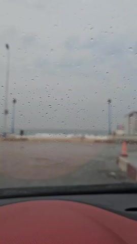The video, viewed from inside a car, shows a beach on a rainy day. Water droplets obscure the windshield. The beach is visible between two light posts.  There is a small building with an "I (heart symbol) (something)" sign on it and an orange cone on the right. The sky is overcast.
