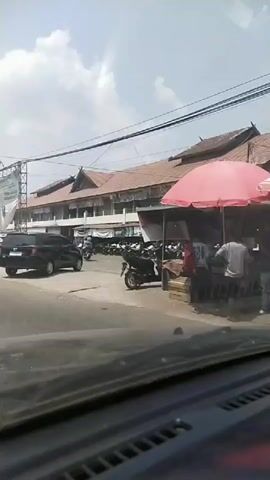 A black car turns left on a paved road. A large banner on a metal frame is visible on the left side of the road. A building with a brown tiled roof and multiple levels is behind the banner. Several motorcycles are parked in front of the building. To the right of the building, a red awning covers a market stall. The sky is cloudy. The video is filmed from inside a vehicle.