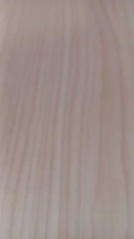 The video begins with a close-up of a light-colored wood grain texture. It then pans to a surface that appears to be a desk or table, with a wall or shelf in the background that has some discoloration. In the next shot, the camera is focused on a stack of papers with colorful elements, and a dark object, possibly a phone, is placed on top. The camera then shifts to show a person wearing a bright red hijab and a matching red robe, seated at a desk and holding papers. The person's face is not clearly visible due to motion blur. The video continues to show the person from different angles, still holding the papers, with a classroom-like environment visible in the background, including desks and what appear to be boards on the wall. The scene suggests an indoor setting, possibly a school or office.