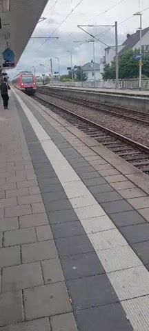 The video shows a train arriving at a station. The train is red and has "DB" written on the front. A person is standing on the platform to the left of the frame, facing the train. The platform is paved with gray and white tiles. The scene is outdoors, with tracks and overhead wires visible. In the background, there are buildings and trees. The sky is cloudy, suggesting it might be daytime. The overall mood appears to be neutral, capturing the arrival of public transportation. There is a sign visible on the platform that reads "630305", likely identifying the train. Another sign further down the platform is partially obscured but appears to be a station name sign.