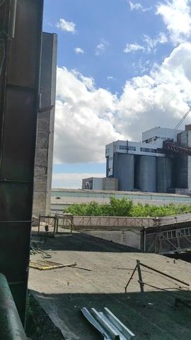 This is a video recording of an industrial complex under a bright, cloudy sky. The camera is positioned at a low angle, looking up towards several tall, concrete silos that are part of a larger building structure. Adjacent to the silos are metal walkways and support beams, forming a complex network of industrial architecture. A crane is visible in the distance.

The scene appears to be an outdoor setting, likely a factory or processing plant, given the presence of industrial equipment and structures. The sky is a vibrant blue with large, puffy white clouds, suggesting fair weather. The sun is shining, casting shadows and illuminating the structures. The time of day appears to be daytime, possibly late morning or early afternoon, based on the lighting. There are no people visible in the recording.

Notable details include the stark contrast between the industrial gray and white structures and the bright blue sky with white clouds. The metalwork of the walkways and supports is intricate and weathered, suggesting age and constant use. The overall impression is one of a functional, operational industrial site. No visible text is present in the video. The location context provided is Kyzyl, Kazakhstan.