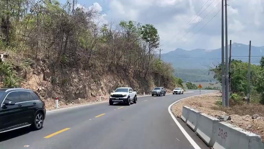 The video shows a road winding through a mountainous, rural area. A white pickup truck passes the camera going in one direction, while a silver pickup truck and a white SUV pass in the opposite direction. There are mountains in the distance and trees on either side of the road. The sky is cloudy. A road sign warns of a sharp turn.