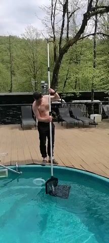The video shows a shirtless man standing on a wooden deck, using a long-handled net to clean a small, round swimming pool. The man is wearing dark pants and shoes. The pool is surrounded by lounge chairs. Behind the deck, there are trees. The sky appears overcast. The man is slowly moving the net through the water.