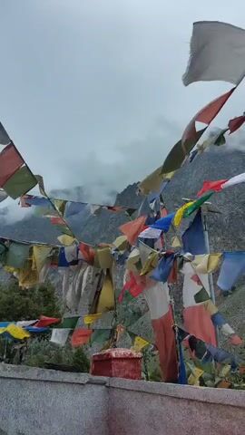 A vertical video displays prayer flags fluttering in the wind against a backdrop of cloudy skies and mountainous terrain. The flags, in various colors and patterns, are strung together and appear to be suspended from poles or wires. The foreground features a weathered concrete wall or structure. The overall scene suggests an outdoor, possibly high-altitude, environment where prayer flags are traditionally used.