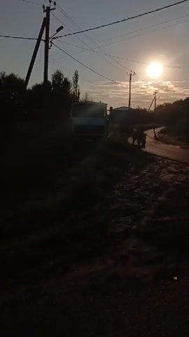 A truck is parked on the side of a road as a person on a tractor drives past. The sun is setting and casts a warm glow over the scene. The road is paved and curves to the right. There are power lines running overhead.