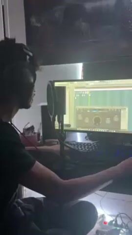 A young man with curly hair is sitting in front of a computer in a dimly lit room. He is wearing headphones and appears to be working with music production software on the computer screen. A microphone and pop filter are positioned in front of him. He is moving his hands and fingers, possibly interacting with the keyboard and mouse or gesturing while listening to the music. Behind him, a framed picture hangs on the wall. The room has some ambient light and a string of small LED lights is visible on the desk.