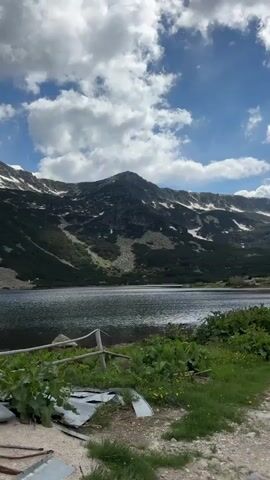 A scenic video pans across a mountain lake. In the foreground, a dirt path runs alongside a grassy bank with some scattered yellow wildflowers. To the left, a section of corrugated metal leans against a wooden fence. Beyond the bank, a large, dark lake stretches across the frame, its surface rippling slightly. In the background, a majestic mountain rises, its slopes covered in a mix of green vegetation and patches of snow. The sky above is a vibrant blue, dotted with fluffy white clouds. The camera's movement is slow and deliberate, offering a sweeping view of the tranquil natural landscape. No people or animals are visible.