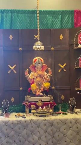 A statue of the Hindu deity Ganesha is displayed on a decorated altar. The statue is seated and adorned with jewelry and a crown. Incense sticks are burning in front of the statue, producing smoke. The altar is covered with a white, patterned cloth and surrounded by decorative items. Behind the altar, a wooden panel backdrop is decorated with gold diamond shapes. Green and pink fabric drapes hang above the backdrop. A decorative metal lamp hangs from the ceiling in front of the statue.