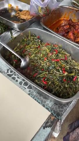 A street food vendor in a market scoops a serving of a vegetable and chili dish into a plastic bag for a customer. The vendor is scooping the food from a large metal tray of stir-fried water spinach with red chilies, which is on a table. Other metal trays of food are visible on the table, including fried chicken and a dish containing flattened fried items. The vendor is wearing a long-sleeved shirt and has light brown skin.