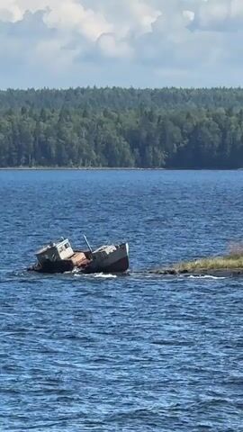 The video shows a shipwrecked boat listing to the side in the water. The boat appears to be an older vessel with visible signs of wear and tear. It is partially submerged and resting against a rocky outcrop or small island. The surrounding environment consists of a large body of water, likely a lake or ocean, with a tree-covered shoreline in the background. The sky above is partly cloudy. The camera is focused on the shipwrecked boat, with the water and distant landscape filling the frame. There are no people visible in the video. The scene conveys a sense of stillness and perhaps abandonment.