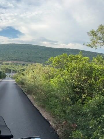 The video shows a view from a moving vehicle, likely a motorcycle, as it travels along a paved road. The road curves to the left and upwards, disappearing over a small hill. On the right side of the road, there is lush green vegetation, with trees and bushes. Beyond the vegetation, there is a valley with more trees and some white buildings. In the distance, there are rolling green hills under a cloudy sky. The perspective suggests the camera is mounted on the front of the vehicle, providing a clear view of the road ahead and the surrounding landscape.