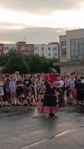 This video captures an outdoor performance in Криводановка, Russia. A group of performers dressed in traditional dark, red-accented clothing are dancing in the street. The performers' outfits feature patterned red trim on their skirts and head coverings. One performer in the foreground is captured mid-spin, their skirt billowing out.

A large crowd of onlookers, of various ages, are gathered to watch the performance. Some people are standing, while others are seated on the wet pavement. The scene appears to be a public event, possibly a festival or community gathering. The presence of a person in a wheelchair suggests an inclusive event.

In the background, several multi-story apartment buildings are visible, indicating an urban or suburban setting. Trees are interspersed between the buildings. The sky is overcast with some lighter clouds, suggesting it might be late afternoon or early evening. The pavement is wet, implying recent rain.

The general atmosphere seems to be one of engagement and enjoyment, with the audience attentively watching the dancers. The clothing of the performers and the style of dance hint at a cultural celebration. There is no visible text in the video that provides specific information about the event.