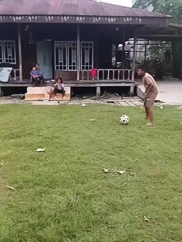 A boy is playing soccer on a grassy field in front of a wooden house. The house has a tin roof and is elevated on stilts. Two women and a child are sitting on the porch of the house. A soccer ball is visible in the grass.