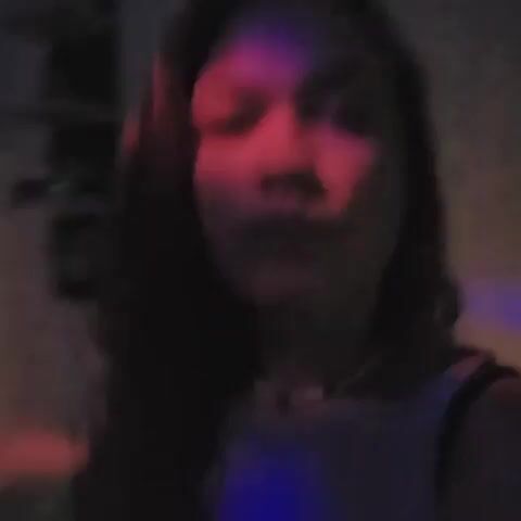 A woman stands indoors with blue and red lights on her face and upper body. The background is dark, but a shelf-like structure is visible. The woman looks directly at the camera. Initially, she appears serious, but she then smiles briefly. She continues to look directly at the camera.
