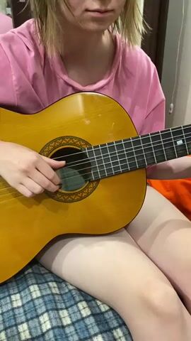 A person wearing a pink t-shirt is playing an acoustic guitar. The person's hands are visible strumming the strings. The guitar is light brown and appears to be an older model. The person is sitting down, and their legs are visible. The background is slightly blurred but appears to be an indoor setting. The person's face is visible, and they appear to be focused on playing the guitar.