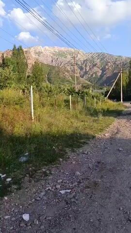 This video depicts a serene, sunlit day in a mountainous region, likely Chimgan, Uzbekistan. The camera pans across a rural landscape, showcasing a gravel road in the foreground that transitions into an asphalt road. A blue car is parked on the side of the road, facing the camera. Beyond the road, a wire fence runs along a grassy area with scattered trees. In the background, imposing, rugged mountains rise majestically under a bright blue sky with scattered white clouds. The light suggests it's late afternoon or early evening.

The overall atmosphere is peaceful and tranquil, characteristic of a rural mountainous setting. The scene appears to be a quiet, undeveloped area, with a few scattered buildings visible in the distance, hinting at a small settlement. The presence of power lines suggests that while rural, the area is connected to essential infrastructure. There is no visible text in the video. The focus is on the natural beauty of the landscape and the simple presence of the vehicle, creating a sense of calm and escape from urban life.