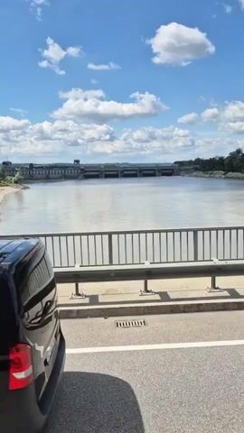 The video shows a view from a bridge over a wide body of water. In the distance, a long, low dam stretches across the water. There is a building or complex structure on the left side of the dam. The water is calm, reflecting the partly cloudy blue sky. The bridge has a metal guardrail and a paved roadway with a white line. The overall scene is an outdoor, daytime landscape.