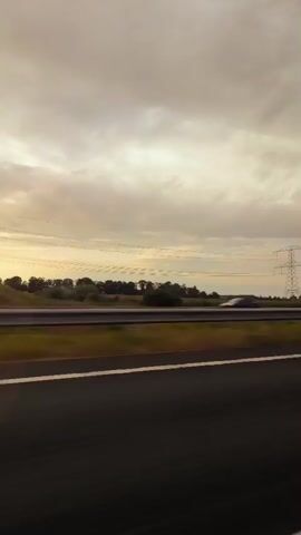The video shows a view from a moving vehicle, likely a car, looking out a window. The scene is dominated by a cloudy sky with a bright horizon, suggesting it's either sunrise or sunset. Below the sky, there is a landscape of trees and fields, with a road running parallel to the vehicle's path. A white van is visible on the road in the distance, moving in the same direction. The perspective is from the side, giving a sense of motion as the scenery blurs slightly. The overall impression is of a journey through a rural or suburban area during twilight.