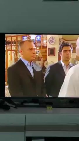 A television displays a scene with three men. Two are dressed in dark suits and stand side-by-side, looking at the third man, who is partially visible with white hair and wearing a white shirt. The background features a display case with decorative items, including plates and vases, and a framed picture on the wall. The television is mounted on a light-colored cabinet with remote controls and a small yellow box on top.