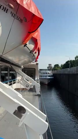 The video shows two ships passing through a lock. The camera is on the deck of the ship in the foreground, looking out towards the second ship. The first ship has a life raft covered with an orange tarp. The second ship is a white passenger vessel with multiple decks. It is moving through a narrow channel between two concrete walls. The sky is clear and blue, and there are trees and a building visible in the distance. The water in the lock appears calm.
